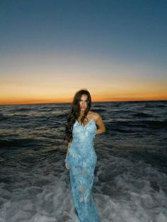 a woman standing in the ocean at sunset wearing a blue dress and holding her hands on her hips