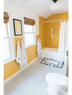 a white toilet sitting next to a bath tub under two windows in a bathroom with yellow walls