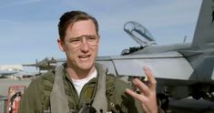 a man wearing glasses standing in front of an airplane with his hand up to the camera