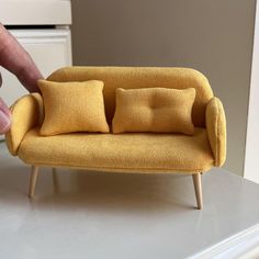 a miniature couch with two pillows sitting on it's back and a hand reaching for the pillow