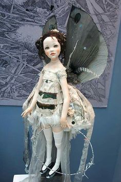 a doll is dressed in white and has wings on her head, as she stands next to a painting