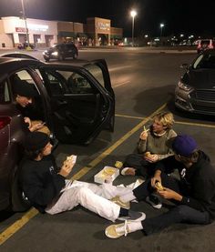 four people sitting in the parking lot eating sandwiches and hotdogs while one person sits on the ground