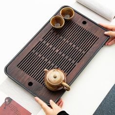 a person holding a teapot on top of a wooden tray next to two cups