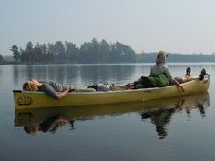 two men and a dog are in a yellow canoe on the water with trees in the background