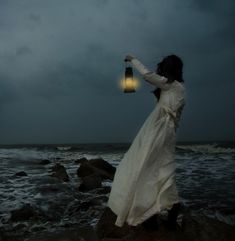 a woman in a white dress is holding a lantern by the ocean on a stormy day