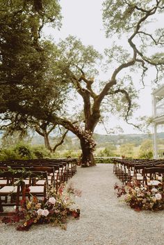 an outdoor ceremony set up with wooden chairs and floral arrangements on the aisle, under a large oak tree