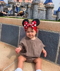 a baby sitting in front of a castle wearing a minnie mouse ears headband and smiling at the camera