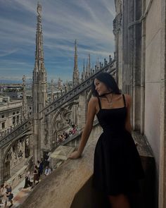 a woman in a black dress is standing on a ledge looking at the city below