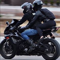 two people riding on the back of a black motorcycle down a street with helmets on