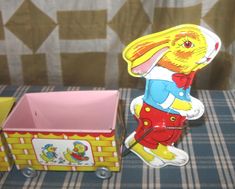 a yellow and red tin toy rabbit next to a box on a plaid tablecloth