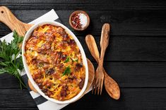 a casserole dish with meat, cheese and parsley next to two wooden spoons