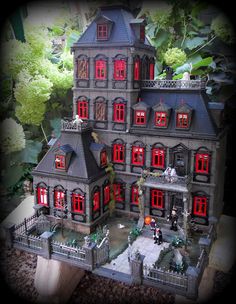 a large doll house with red windows and lights on the front, surrounded by greenery