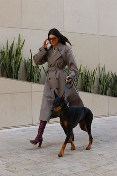 a woman in a trench coat walking her dog on a leash and talking on the phone