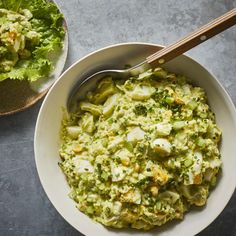 a white bowl filled with food next to a salad on top of a plate and a wooden spoon