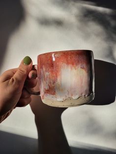 a hand holding a coffee cup with red and white paint on the outside, in front of a window