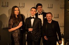 three men and two women standing next to each other in front of a red carpet