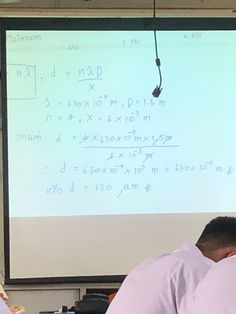 two men sitting at desks in front of a projector screen with writing on it