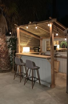 an outdoor bar with two stools and lights on the outside wall, next to a palm tree