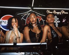 three beautiful women standing next to each other in front of a neon sign at night