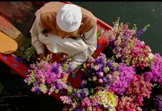 a man sitting in a boat filled with lots of purple and white flowers next to water