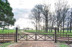 a gated dirt road leading to a grassy field with trees in the back ground