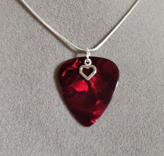 a red guitar pick with a heart charm on a white cord necklace hanging from a silver chain