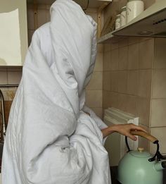 a person wrapped in a blanket is cooking on the stove top with a tea kettle