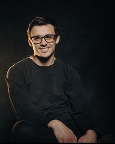 a man wearing glasses sitting in front of a black background with his hands crossed and smiling at the camera