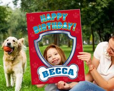 a woman sitting in the grass holding up a birthday card with a dog behind her