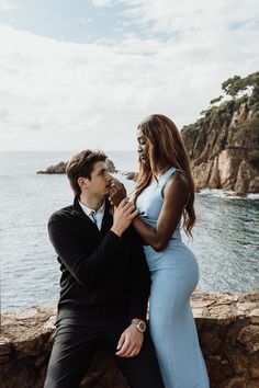 a man and woman sitting next to each other near the ocean with an ice cream cone in their mouth