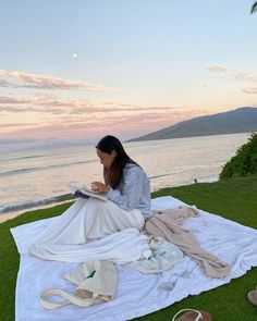 a woman sitting on top of a white blanket next to the ocean reading a book