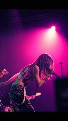 two young women on stage playing guitars and singing into microphones with bright lights behind them