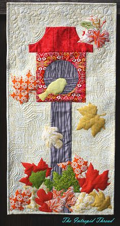 a quilted wall hanging with a birdhouse and flowers on it's side