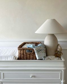 a white dresser with a basket on top of it next to a lamp and stuffed animals