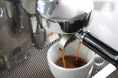 espresso being poured into a white cup with saucer on the side and coffee machine in background