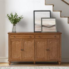 a wooden sideboard with two framed pictures on top and a plant in the corner