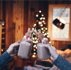 two people holding up mason jars with marshmallows in front of a christmas tree