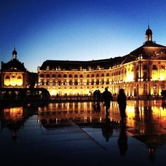 people are walking in front of a large building at night with its lights on and reflecting off the wet ground