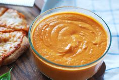 a small glass bowl filled with sauce next to a pizza on a wooden cutting board