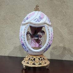 an ornate white and gold egg with pink flowers in it's center sits on a wooden table