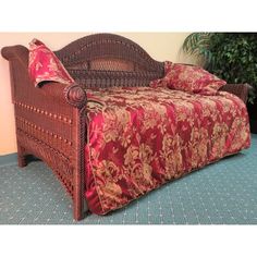 a wicker bed with red and gold bedspread on it in front of a potted plant