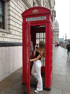 a woman in white pants is standing inside a red phone booth on the sidewalk near a building