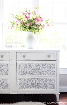 a white dresser with pink flowers in a vase sitting on top of it next to a window