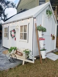 a small white shed with potted plants on the side and a wheelbarrow next to it