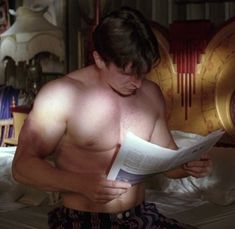 a shirtless man reading a paper while sitting on a bed