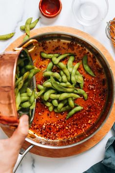 someone is pouring sauce into a pot with green beans in it and another person's hand holding a spoon