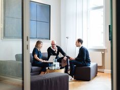 three people sitting on couches in a living room, one is holding a laptop