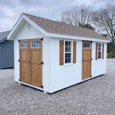 a small white shed with brown shutters and windows on gravel ground next to trees