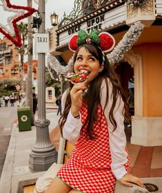 a woman in red and white dress sitting on the curb eating food with minnie ears