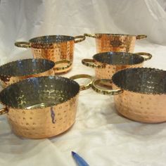 six copper colored pots with handles sitting on a white cloth next to a blue pen